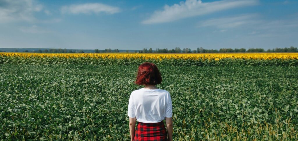 Eine junge Person mit roten Haaren und einem schwarz-rot karierten Hemd, das um die Hüfte gebunden ist, steht mit dem Rücken zur Betrachterin vor einem Acker.