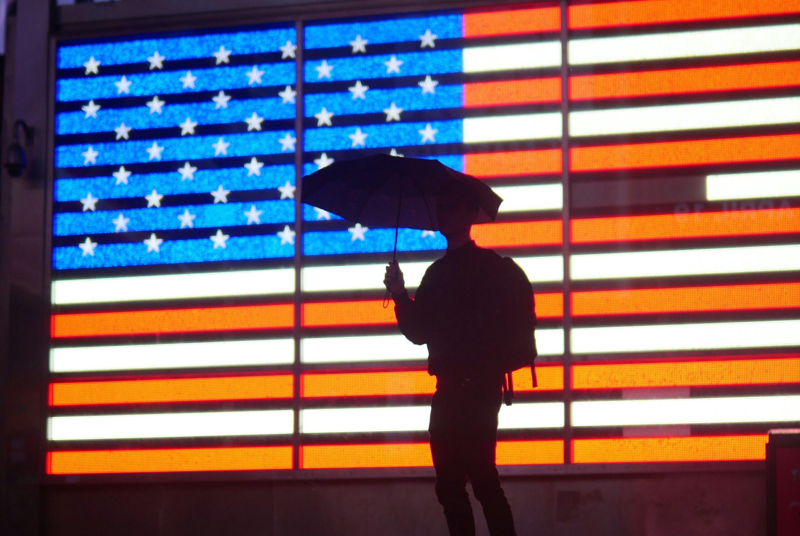 Silhouette einer Person mit einem Regenschirm, die vor einer Leuchttafel mit der US-Flagge steht.