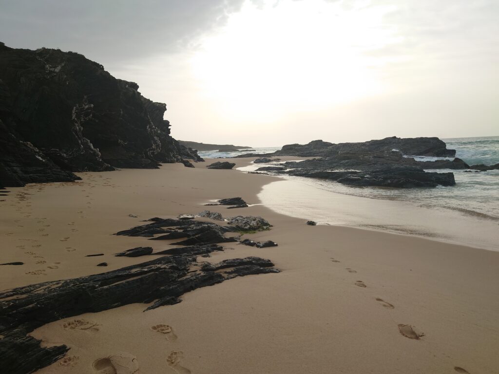 Ein Strand mit rauen Felsen und einem wolkigen Himmel, hinter dem die Sonne leuchtet.