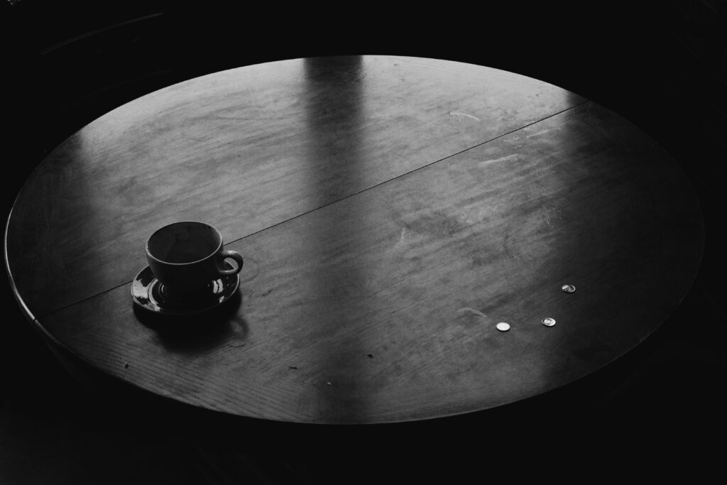 Runder dunkler Tisch mit einer Kaffeetasse auf Untertasse; rechts liegen einige Münzen. Aufnahme in Schwarz-Weiß.