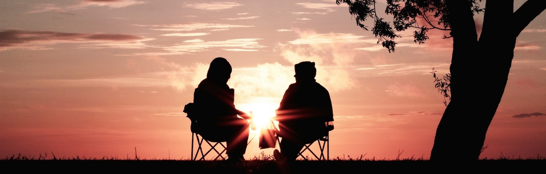 Zwei Personen sitzen im Sonnenuntergang unter einem Baum und blicken gemeinsam in den farbigen Abendhimmel.